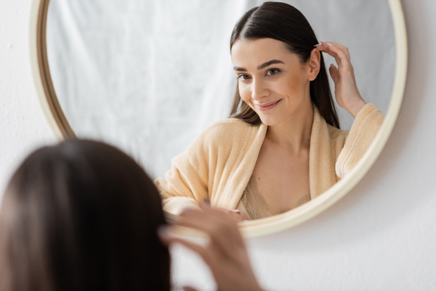 young brunette woman adjusting hair and smiling while looking at mirror in bathroom.
