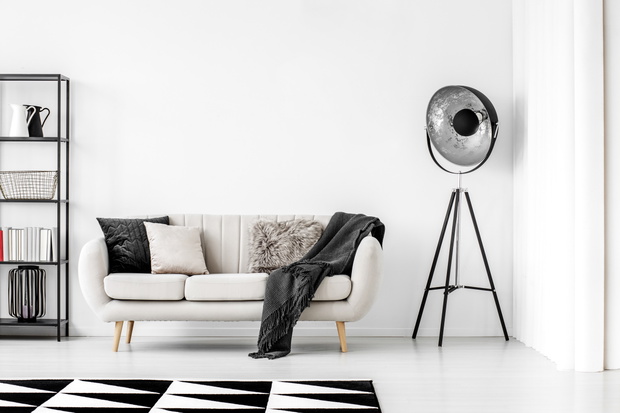 Industrial black lamp next to beige couch with blanket and pillows, copy space on empty white wall