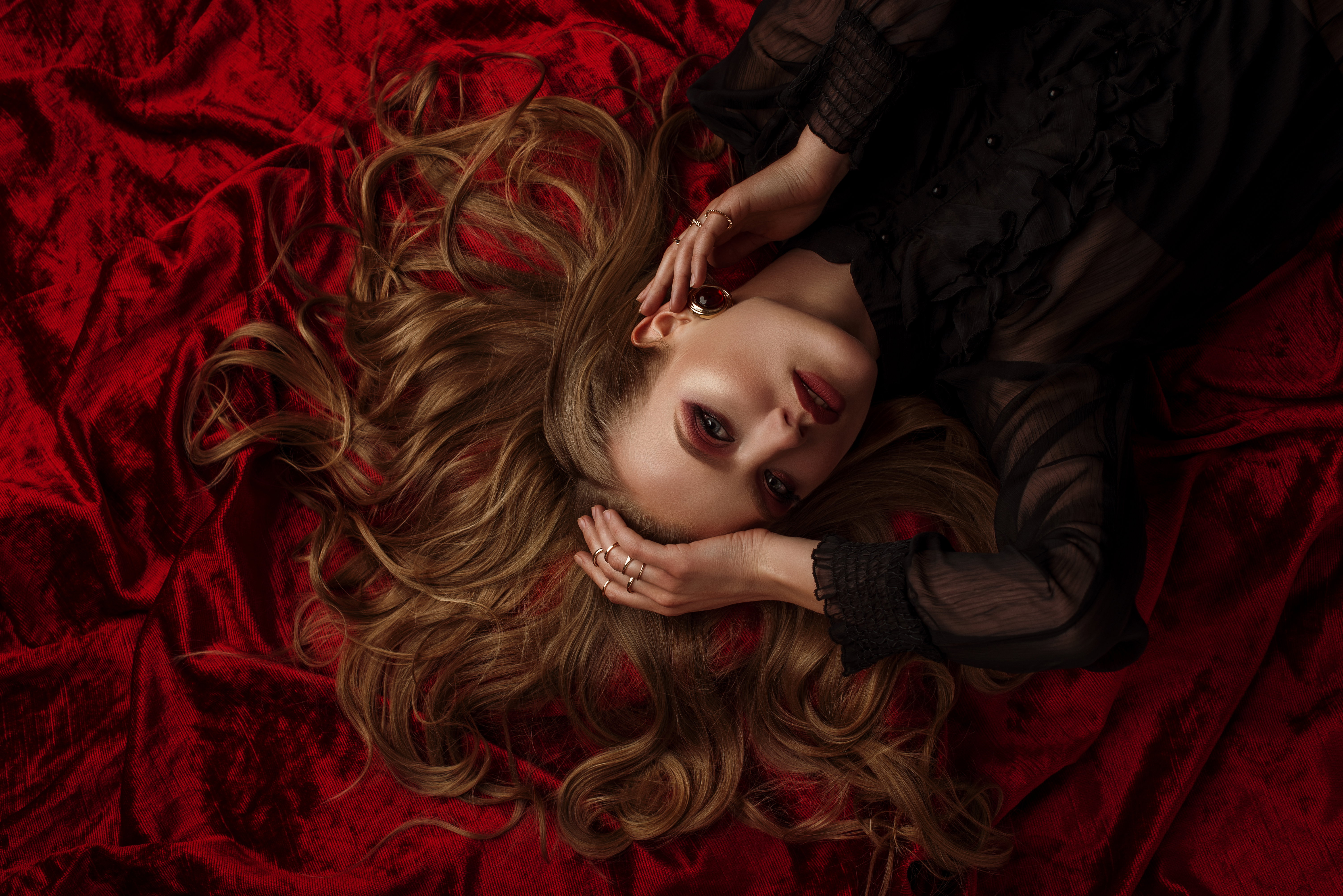 Beautiful lady with luxury blond hair, glow skin, dark red eyes and lips makeup, wearing a lot of golden rings, stylish earrings, posing, laying on velvet sheets. Close up studio portrait