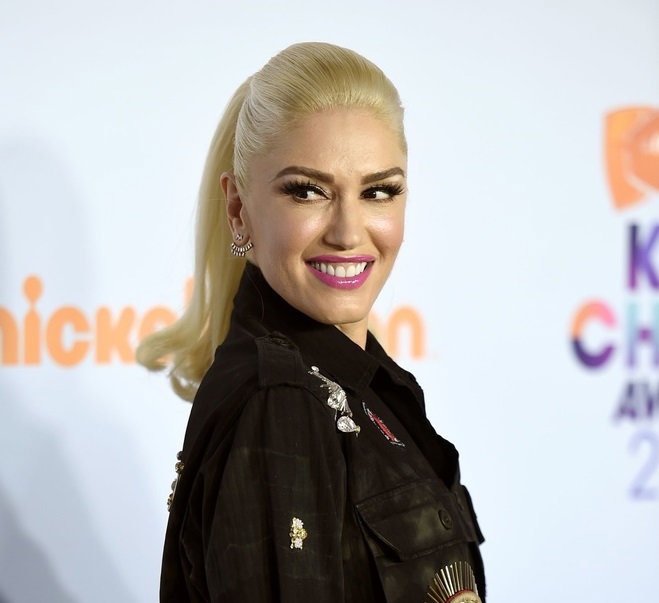 Gwen Stefani w blond włosach związanych w oszukanego uśmiecha pozuje na niebieskim tle w czarnej kurtce