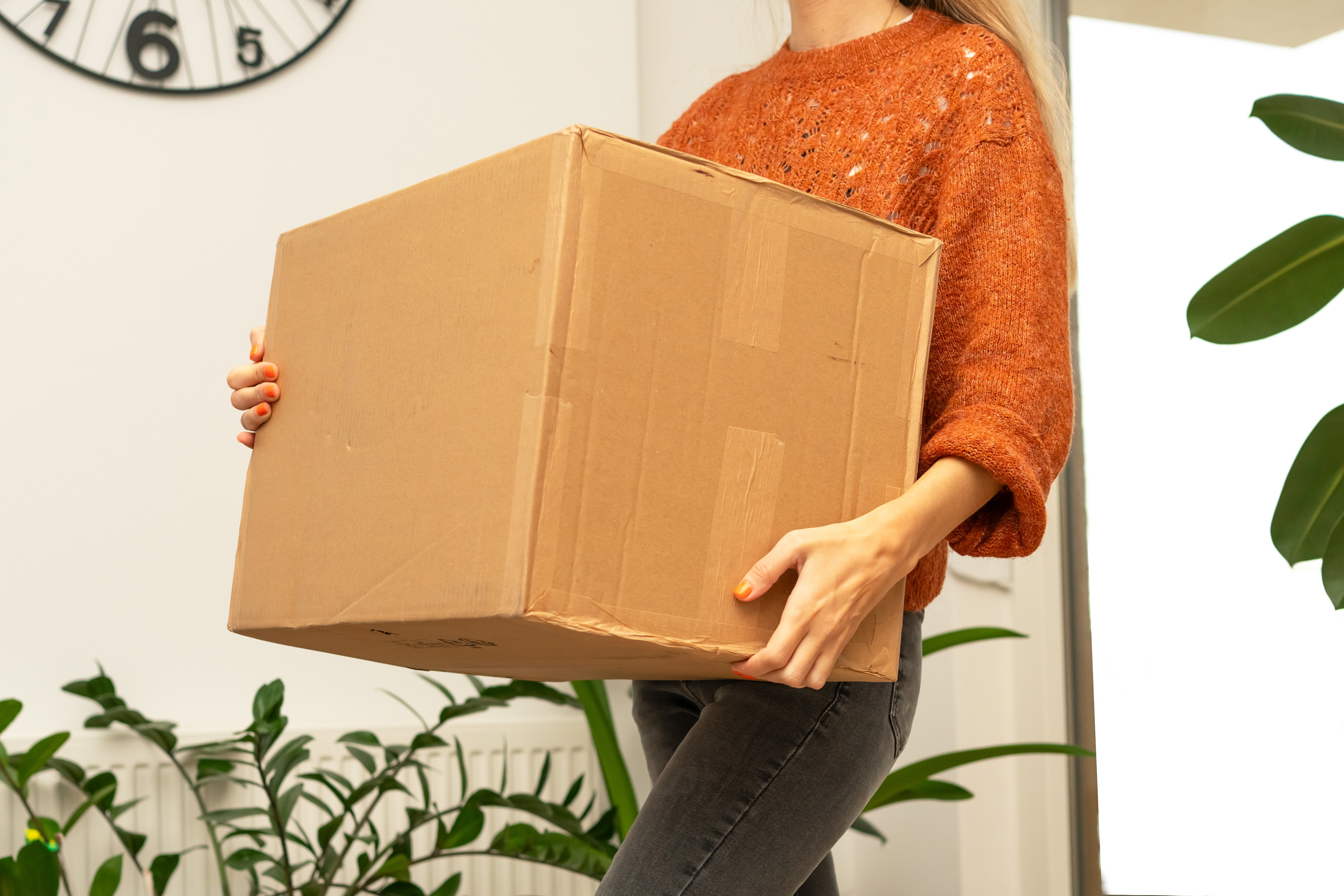 A young woman at home in a room with green plants holds a large cardboard box in her hands. Concept of delivery, online shopping, sending gifts. High quality photo