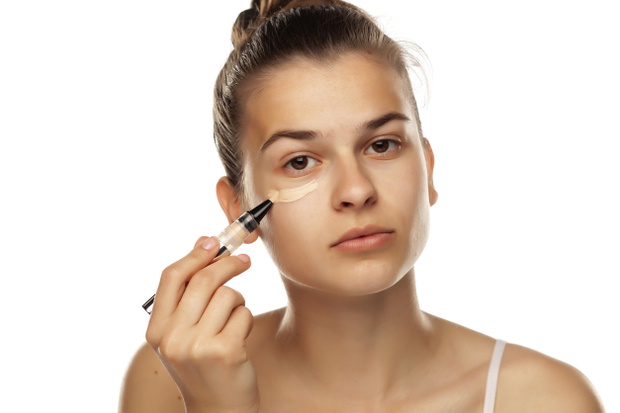 Portrait of young woman applying concealer with brush on white background