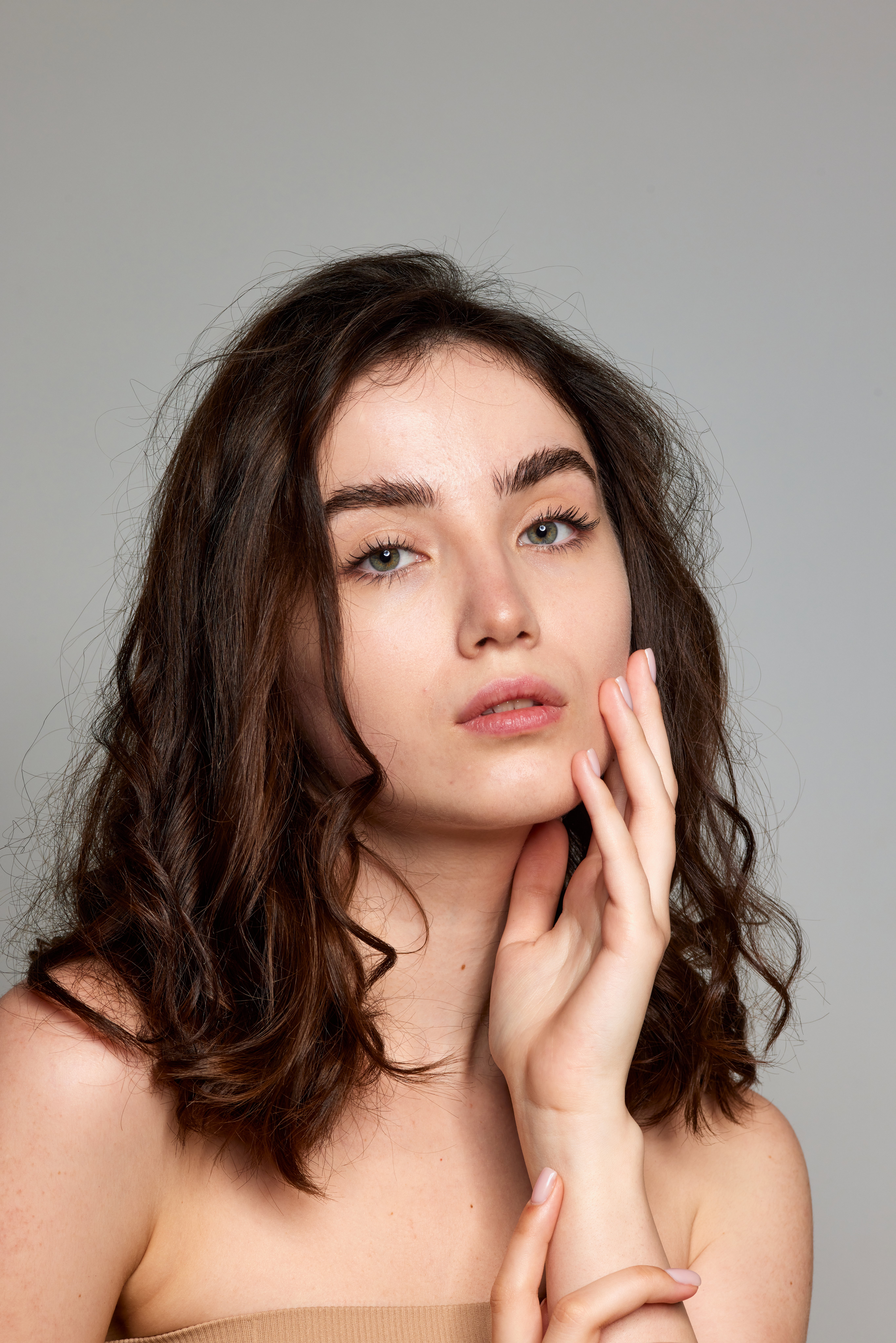 Curly hair. Tender look. Portrait of young beautiful brunette girl with well-kept skin over grey studio background. Concept of natural beauty, skincare, cosmetology