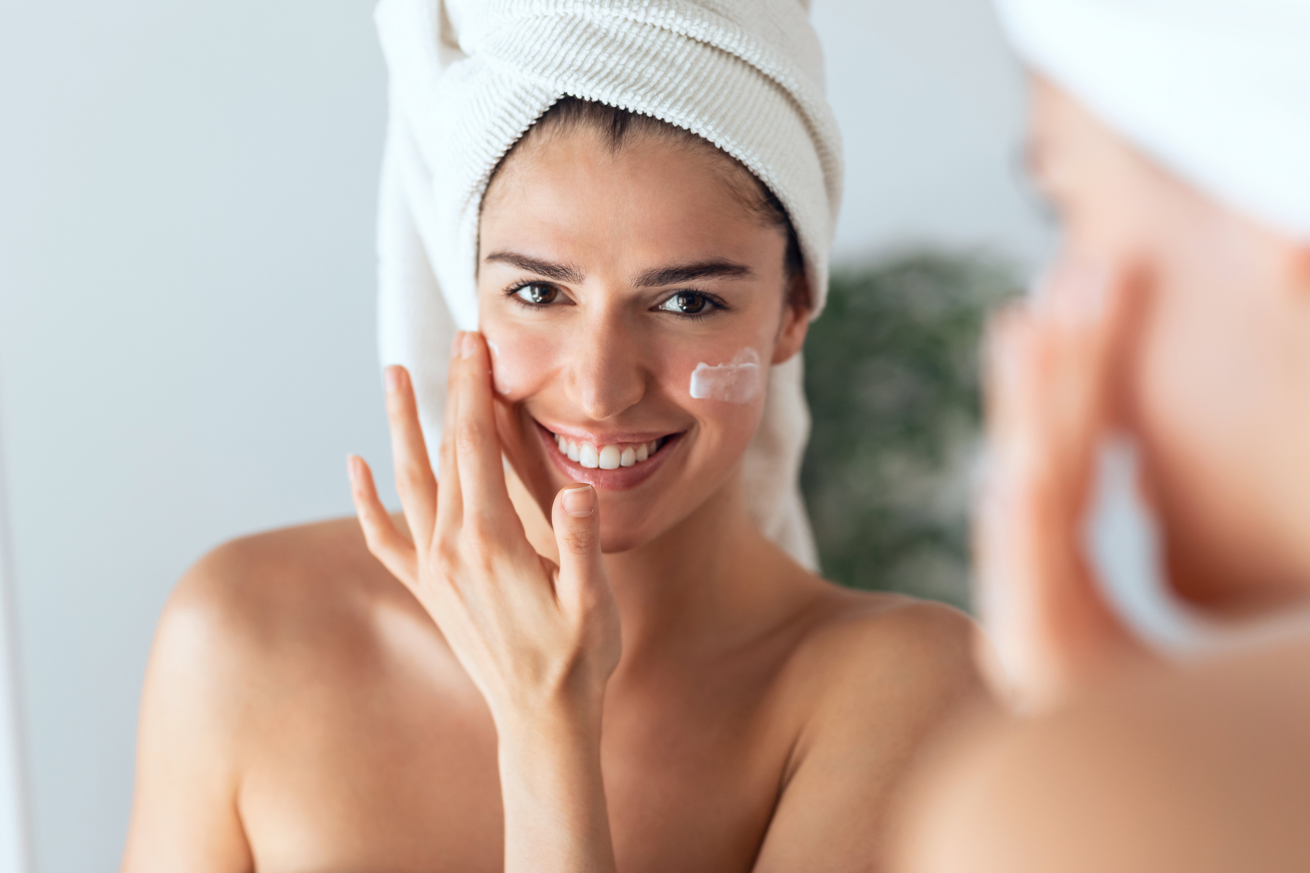 Beautiful young woman caring of her skin while putting on cream looking at mirror at home.