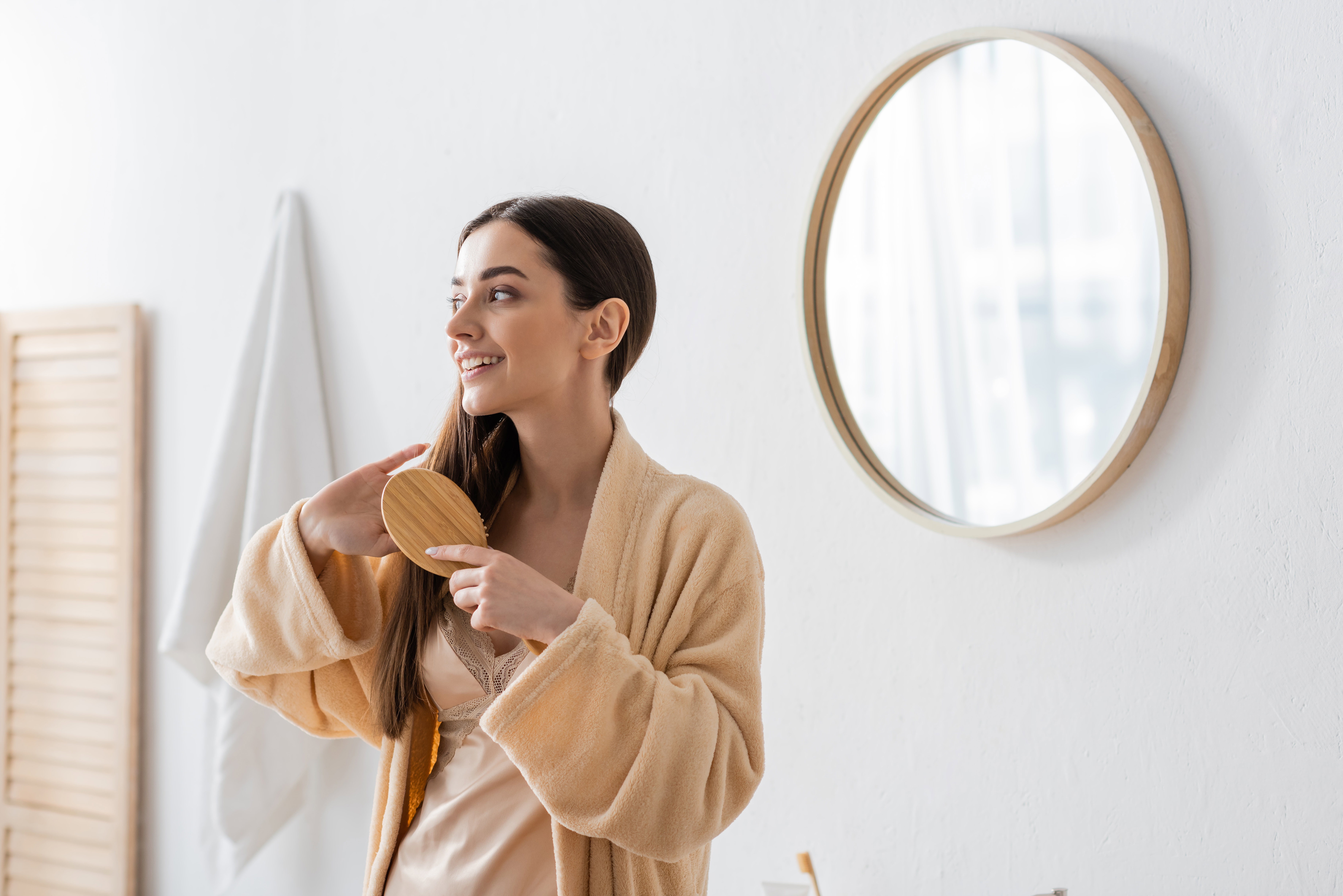 happy young woman in bathrobe brushing hair in white modern bathroom.